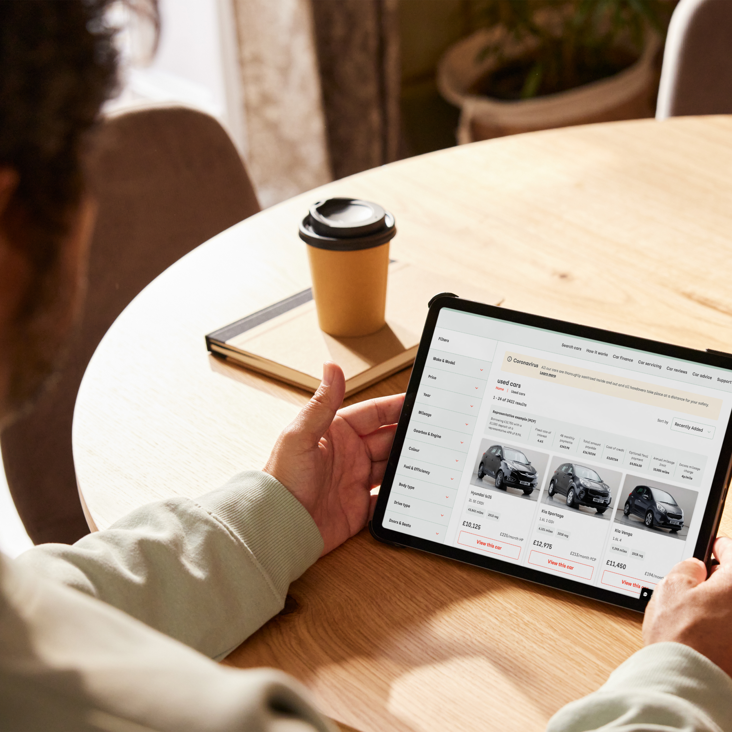 A person is sitting at a wooden table, holding a tablet while browsing for used cars online. 