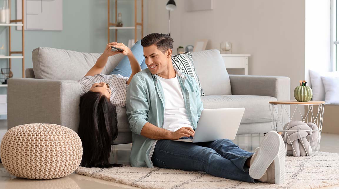 Young caucasian couple sitting on their grey couch checking their mobile phone and silver laptop