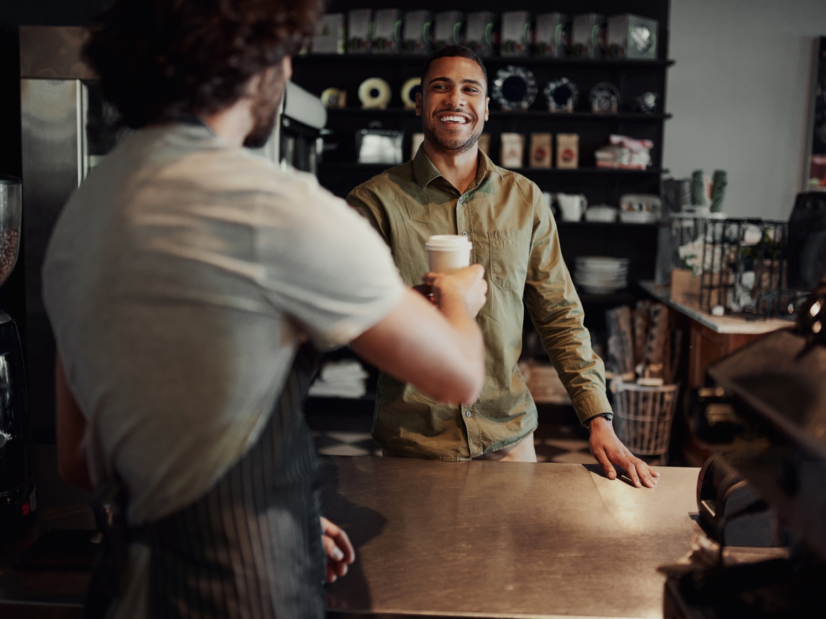 A customer at a café counter smiles as they receive a takeaway coffee cup from a barista. The customer, dressed in a light green button-up shirt, stands on the other side of the counter. The café background features shelves with various items, such as mugs and coffee bags. The atmosphere is warm and inviting.