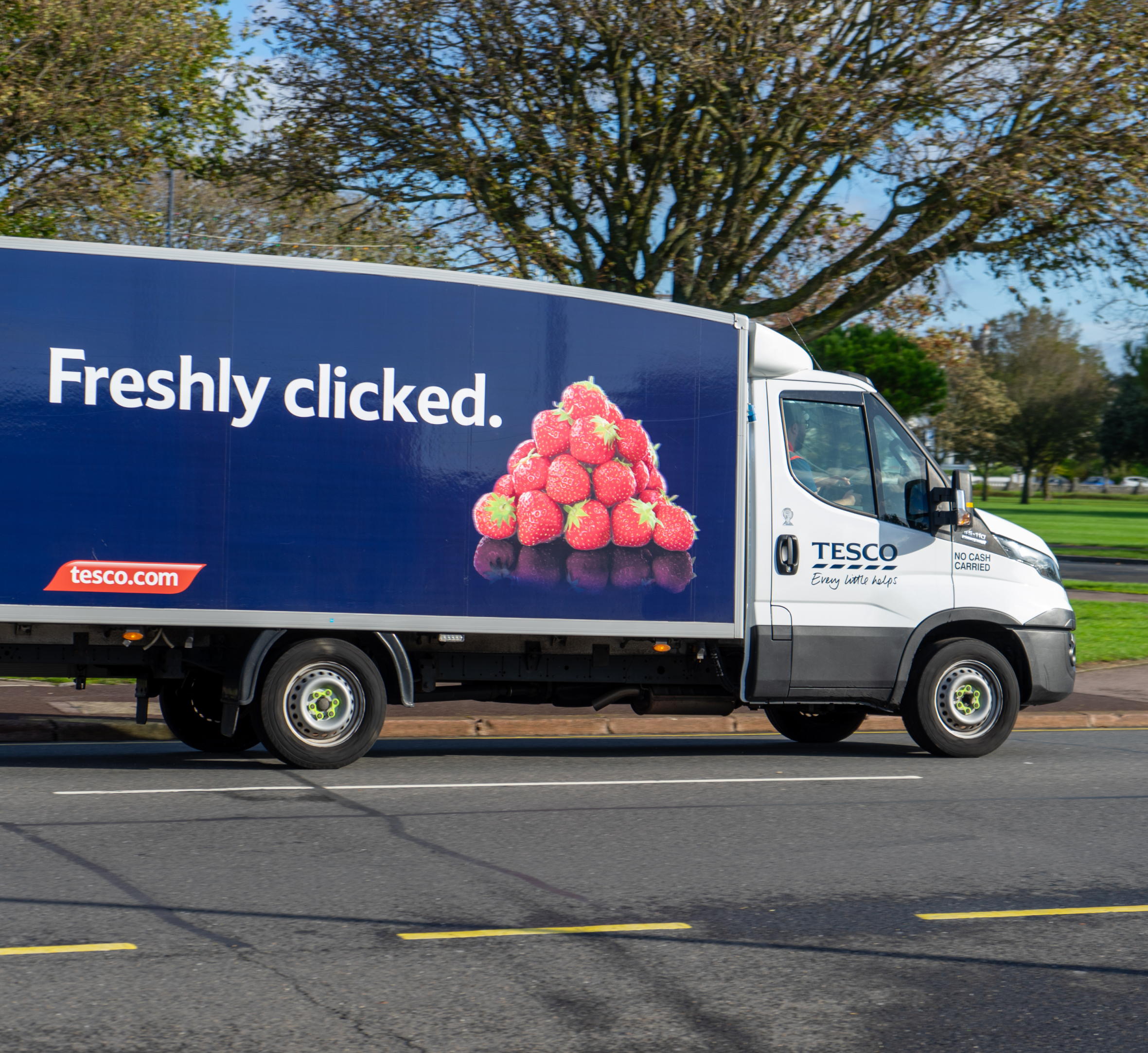  "A Tesco delivery truck driving on a road, featuring an advertisement on the side that reads 'Freshly clicked' with an image of fresh strawberries. The truck promotes Tesco's online grocery service, with a logo and the website 'tesco.com' displayed on the side.