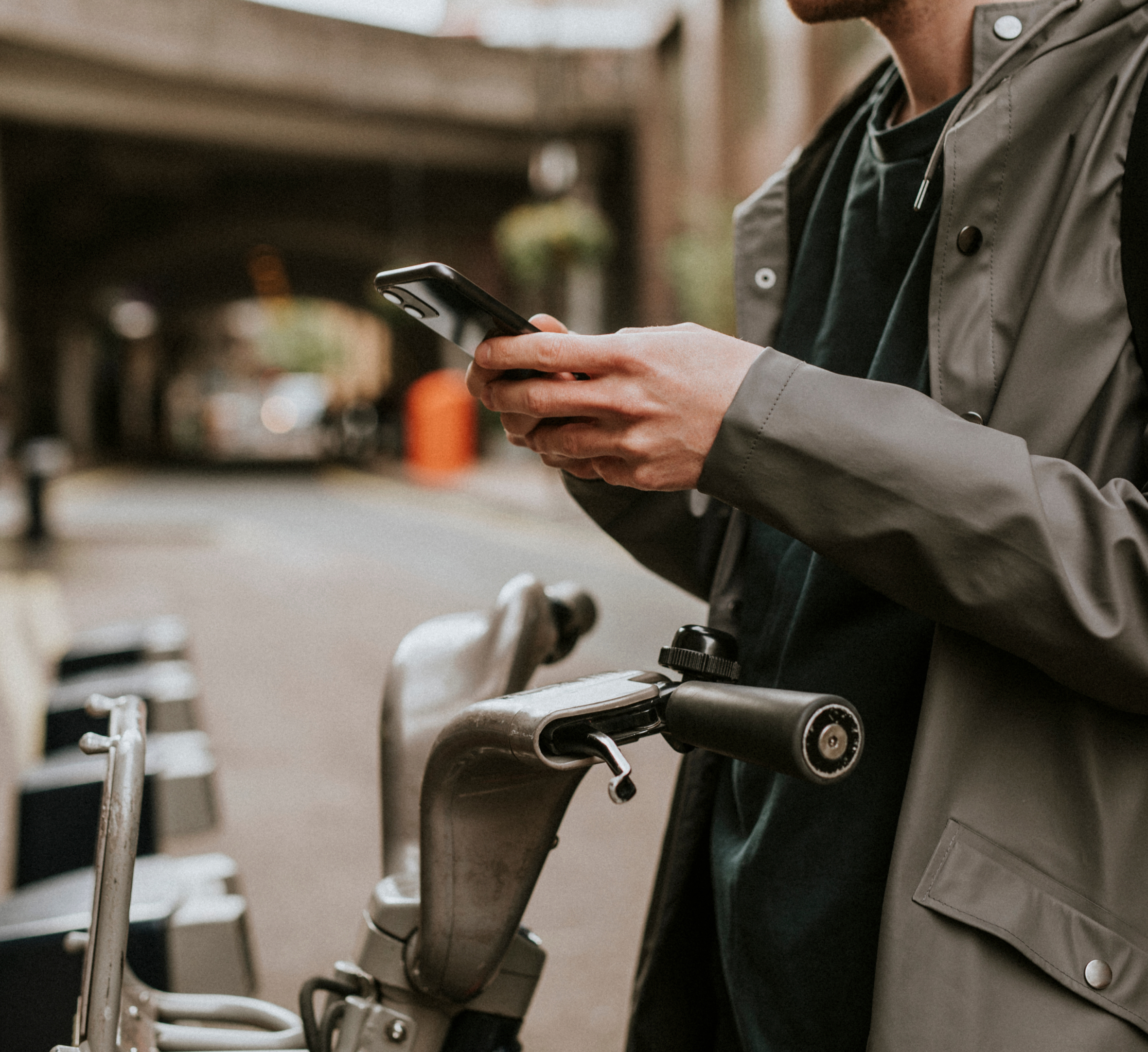 A person standing next to a bike, holding a smartphone and checking something on the screen. The individual is dressed in a raincoat, with the scene taking place on a street near a bridge, capturing an urban cycling moment.