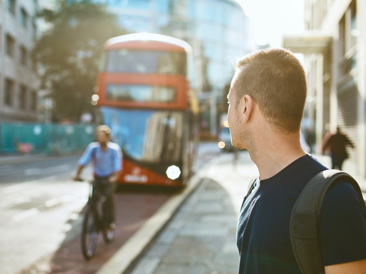 A man with a backpack stands on a city street, looking toward an approaching red double-decker bus. A cyclist rides past on the street, with blurred city buildings in the background, suggesting an urban commuting scene.