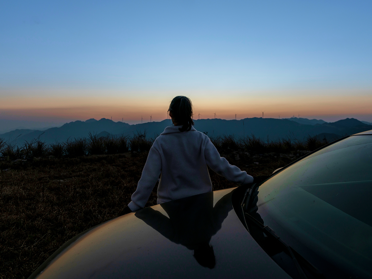 A woman stands beside a car, silhouetted against a vibrant sunset sky.