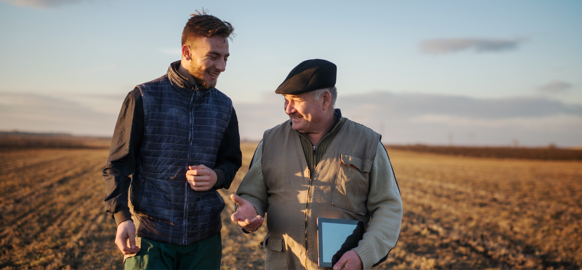 two farmers talking and walking in a field at sunset, one holding a tablet