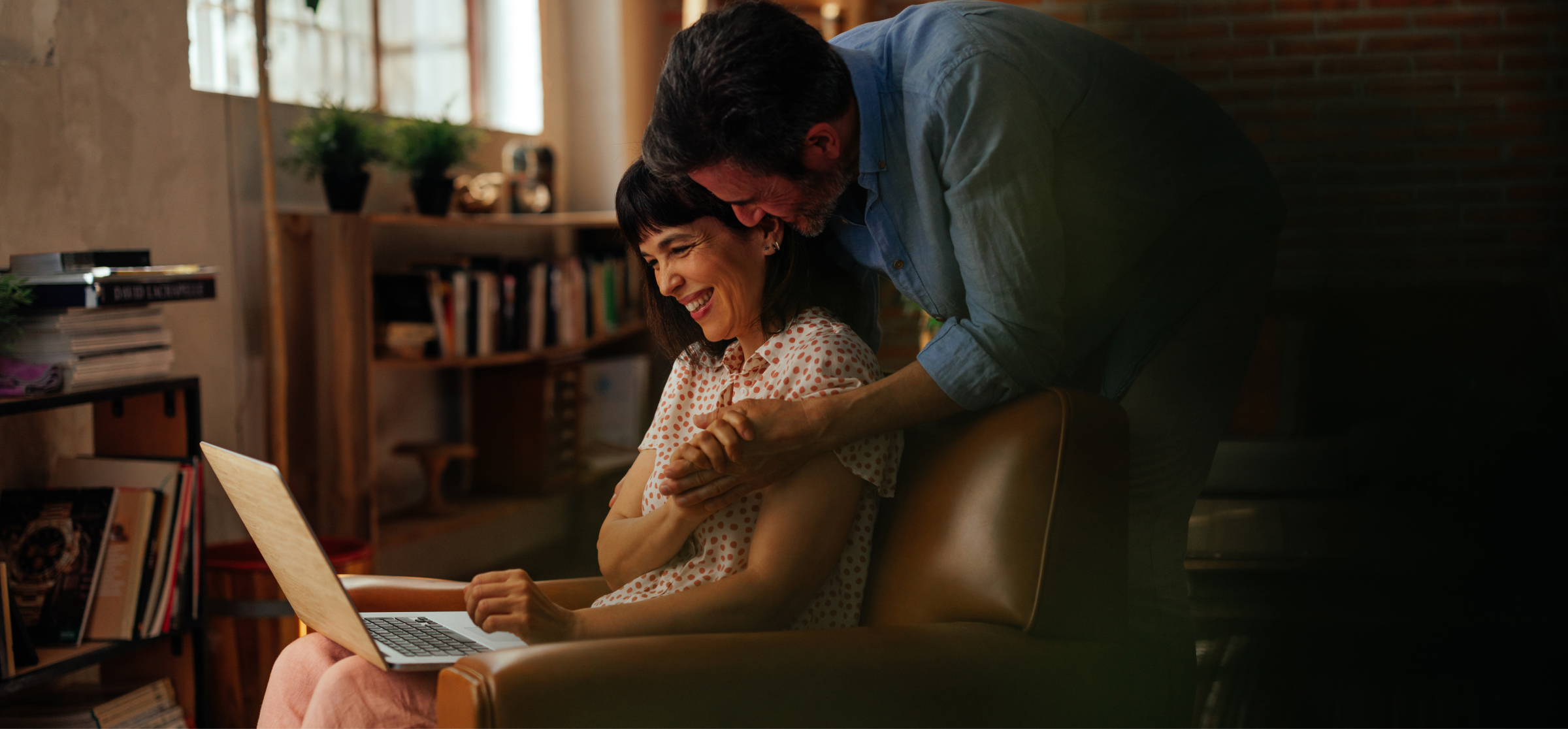 A couple, with the man leaning over the woman’s chair, share a joyful moment while looking at a laptop. The woman is seated in a cozy living room filled with bookshelves and plants, smiling as they browse something on the screen together. The warm lighting and relaxed setting suggest a peaceful and happy atmosphere.