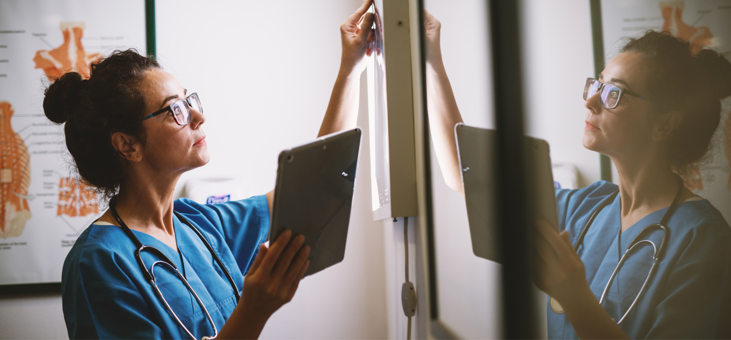 A female healthcare professional, wearing blue scrubs and a stethoscope, is carefully examining an X-ray on a lightbox while holding a tablet in her other hand. She is focused on the X-ray, studying it closely, with her reflection visible on the glass surface next to her. Behind her is a medical chart displaying anatomical diagrams. The image captures the use of both traditional and digital tools in medical diagnostics, emphasizing precision and attention to detail in healthcare settings.