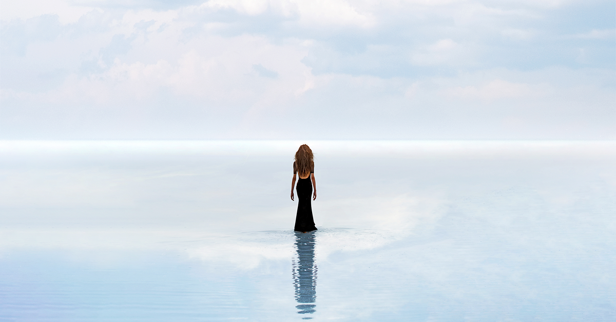 Elegant woman in a black dress standing in shallow water, gazing toward the serene horizon under a pastel sky, evoking luxury and tranquility.