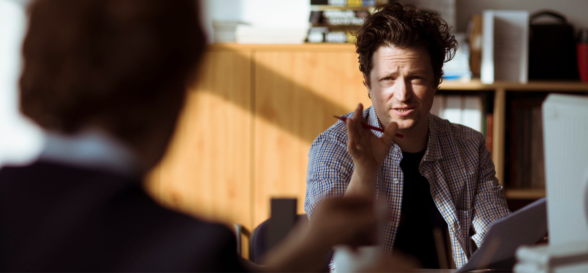 A man with curly hair is engaged in a discussion, holding a pen in his hand while speaking. He is wearing a checkered shirt and sitting in an office environment, with shelves of books and documents behind him. Sunlight is streaming into the room, casting soft shadows, creating a warm and professional atmosphere. The man appears focused, gesturing as he talks, likely in the middle of a meeting or collaborative discussion. The image emphasizes communication and engagement in a work setting.