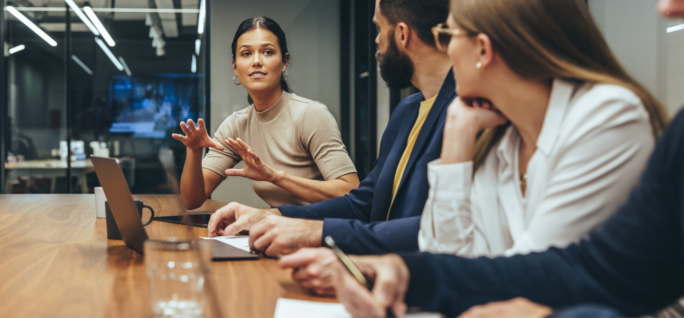 A woman is actively speaking and gesturing with her hands during a meeting, engaging her colleagues in a discussion. She is seated at a conference table with two other people, one of whom is taking notes. The group is dressed in business attire, and the setting is a modern office with glass walls and a large screen in the background. The woman appears confident and expressive, leading the conversation. The image highlights teamwork, communication, and collaboration in a professional environment.