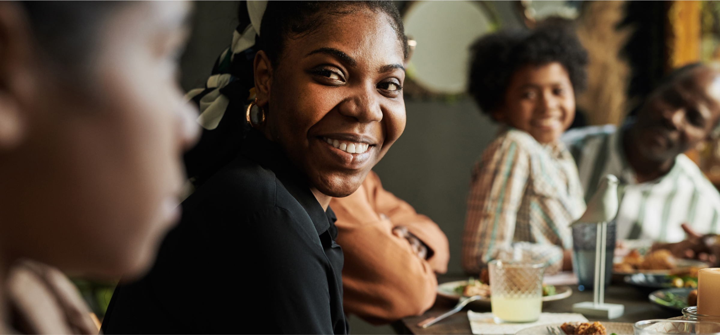 A young woman is smiling warmly at the camera while sitting at a dinner table with her family. She is wearing a black shirt and gold earrings, and her hair is tied back with a patterned scarf. In the background, other family members, including a young boy and an older man, are also smiling and enjoying the meal. The table is set with plates of food, drinks, and a candle, creating a cozy and welcoming atmosphere. The image captures the joy of shared moments and connection during a family gathering.