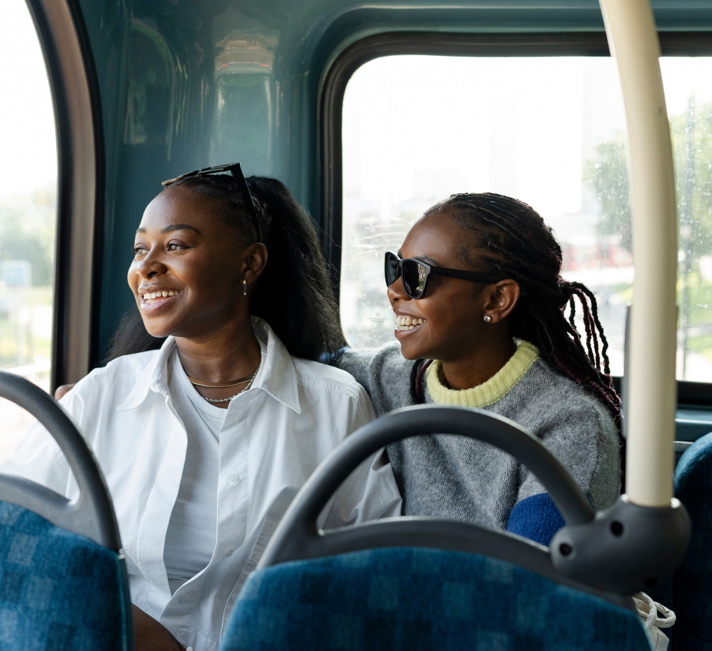 Two women are sitting together on a bus, smiling and enjoying each other’s company. One woman, wearing a white shirt and sunglasses on her head, looks out the window with a bright smile. The other woman, wearing a gray sweater and dark sunglasses, has her arm around her friend and is laughing. They appear to be having a joyful moment during their journey. The background shows the sunlight filtering through the bus windows, creating a warm and pleasant atmosphere. The image captures a happy, relaxed moment between friends on public transport.