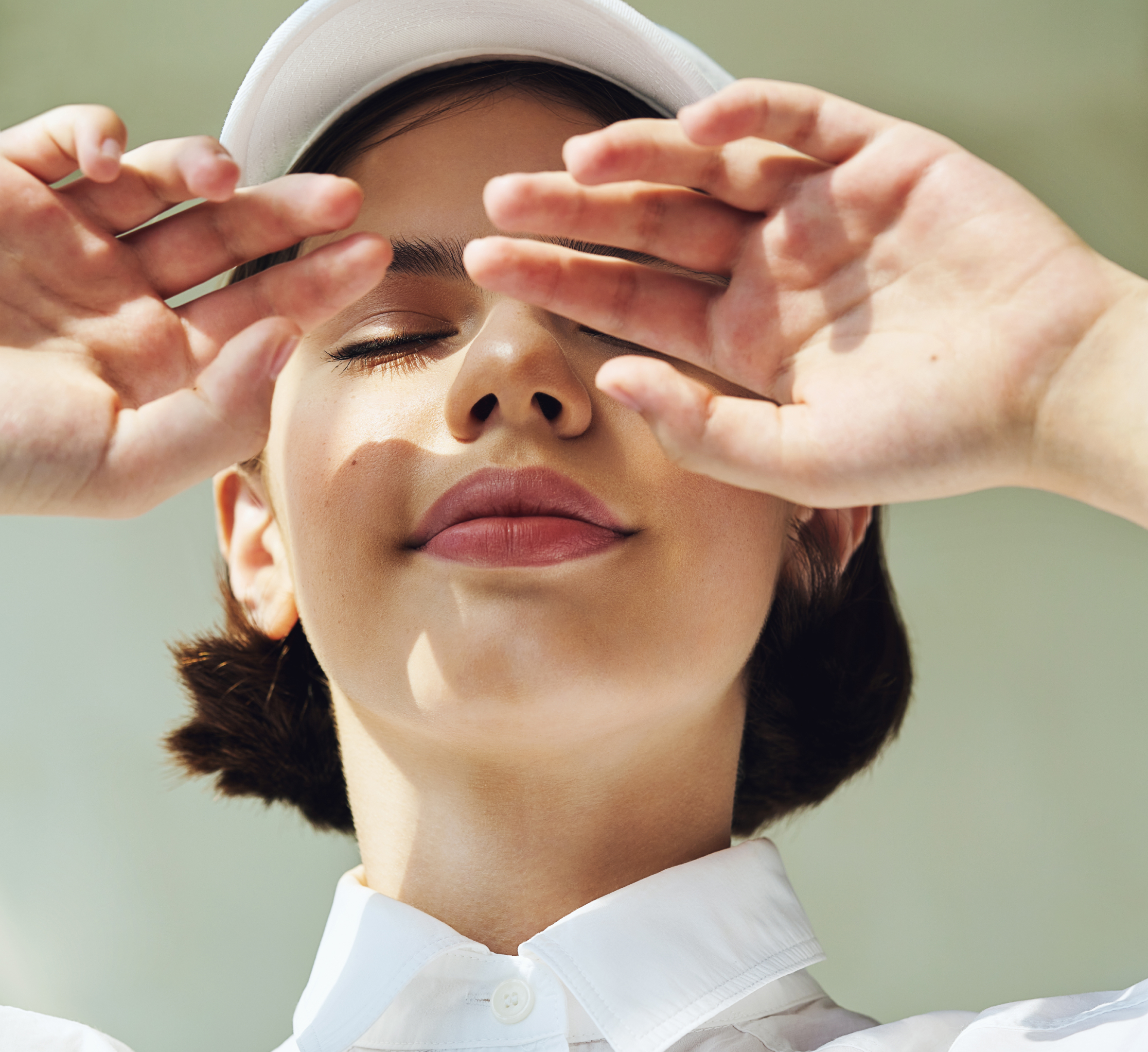 A woman in a white cap with her eyes closed, smiling gently while raising her hands to her face, blocking the sunlight.