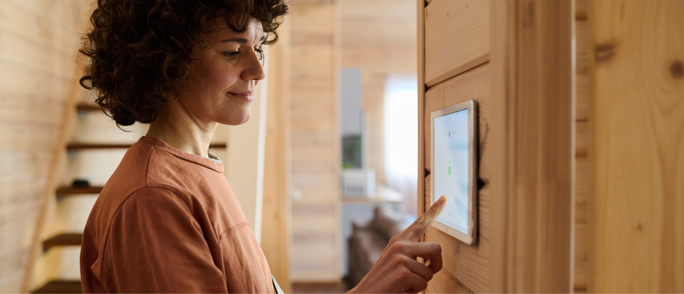 A woman interacting with a touch-screen control panel mounted on a wooden wall inside a home.