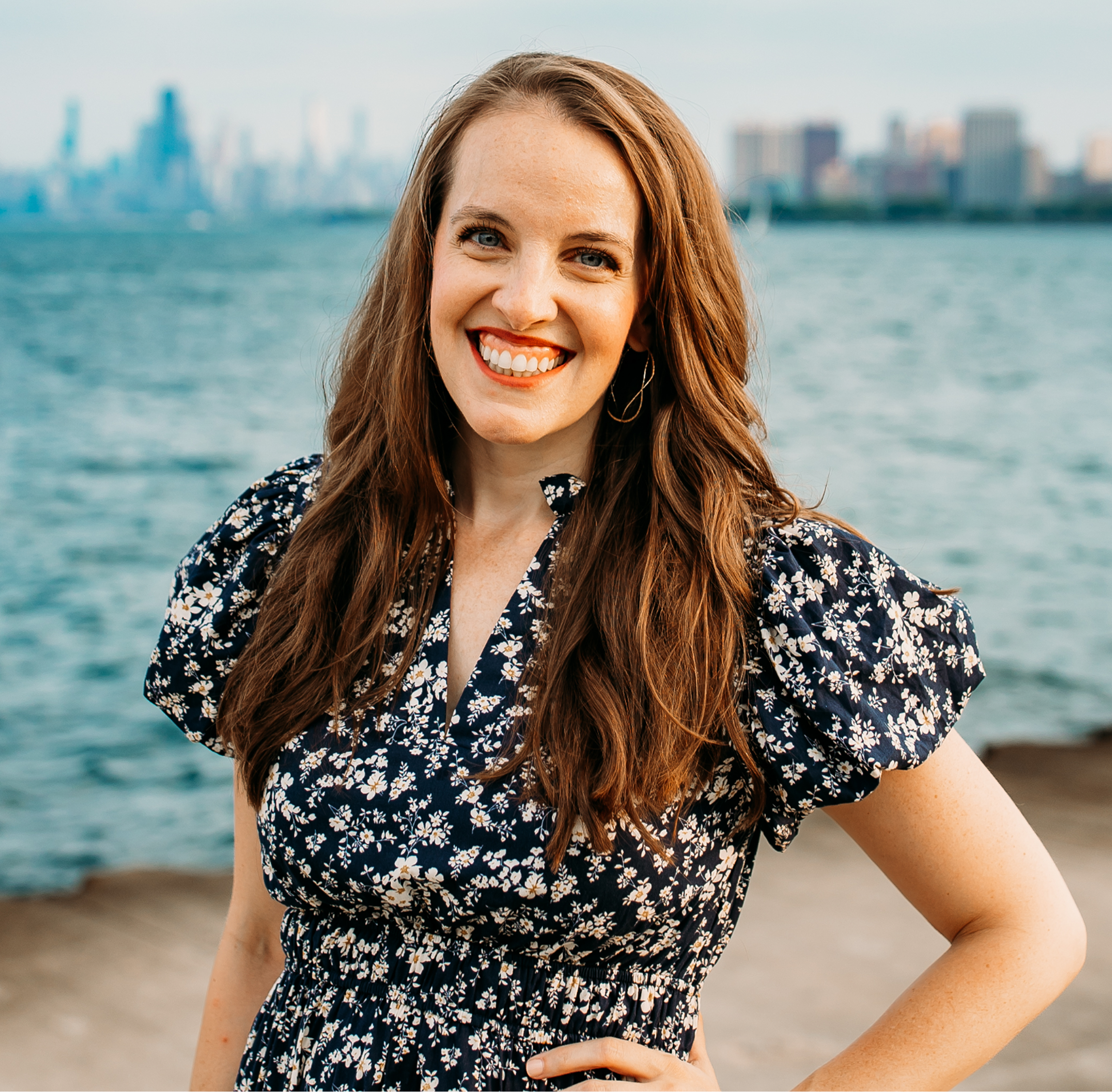 Kerry Penny, SVP and Global Head of Brand & Content at Valtech, stands near a waterfront with a blurred city skyline in the background. She is wearing a navy blue floral dress with puffed sleeves and gold hoop earrings. The weather appears clear, and the water behind her is calm.