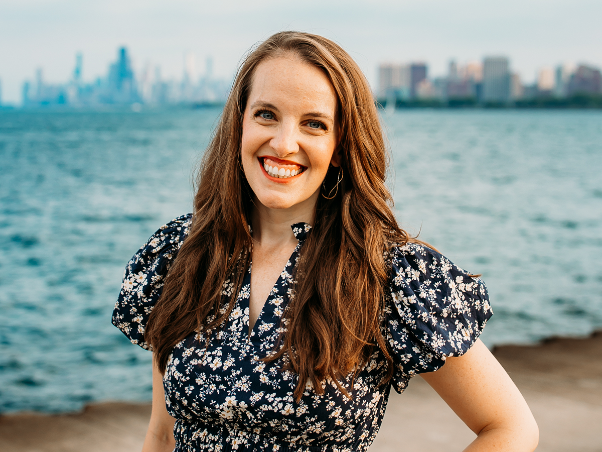 Kerry Penny, SVP and Global Head of Brand & Content at Valtech, stands near a waterfront with a blurred city skyline in the background. She is wearing a navy blue floral dress with puffed sleeves and gold hoop earrings. The weather appears clear, and the water behind her is calm.
