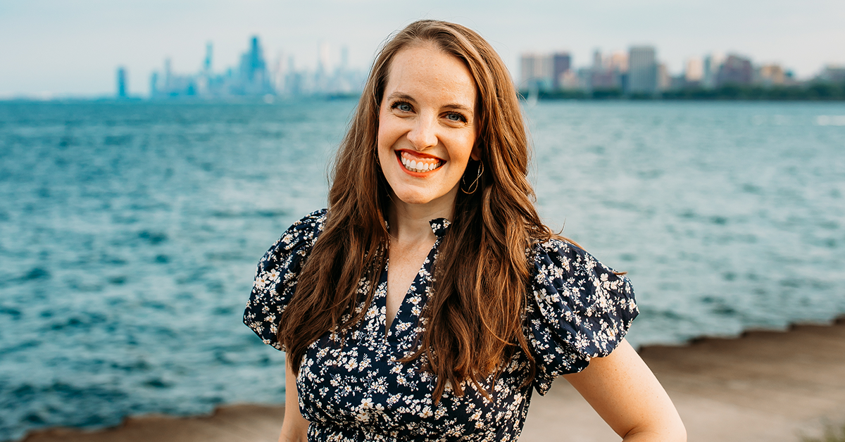Kerry Penny, SVP and Global Head of Brand & Content at Valtech, stands near a waterfront with a blurred city skyline in the background. She is wearing a navy blue floral dress with puffed sleeves and gold hoop earrings. The weather appears clear, and the water behind her is calm.
