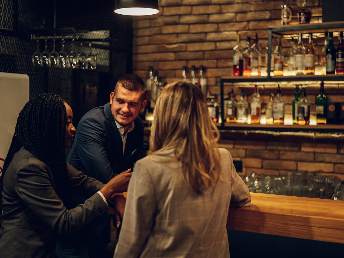 Three business professionals—two women and one man—chatting at a stylish bar counter with exposed brick walls and backlit liquor shelves.