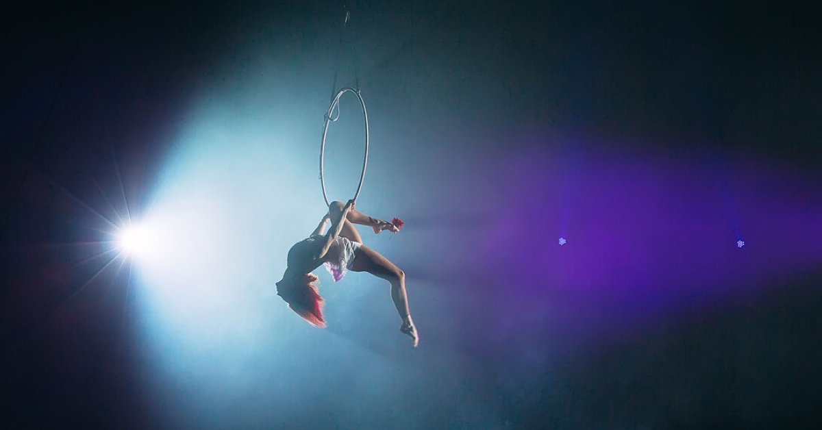 Aerial acrobat performing on a suspended hoop under dramatic stage lighting with blue and purple hues. The performer is mid-movement, displaying strength and elegance in a gravity-defying pose.