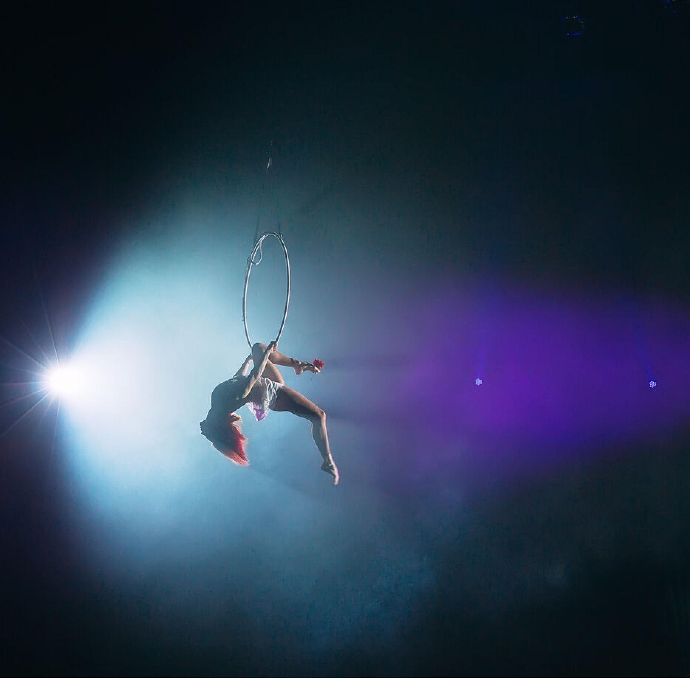 Aerial acrobat performing on a suspended hoop under dramatic stage lighting with blue and purple hues. The performer is mid-movement, displaying strength and elegance in a gravity-defying pose.