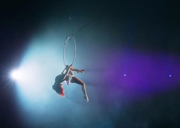 Aerial acrobat performing on a suspended hoop under dramatic stage lighting with blue and purple hues. The performer is mid-movement, displaying strength and elegance in a gravity-defying pose.