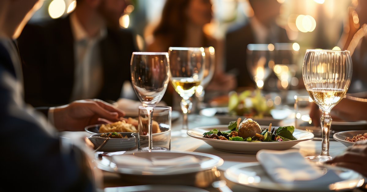 Close-up of a sophisticated dinner table during a business event, featuring wine glasses, gourmet dishes, and elegantly dressed people engaged in conversation in a warmly lit setting.