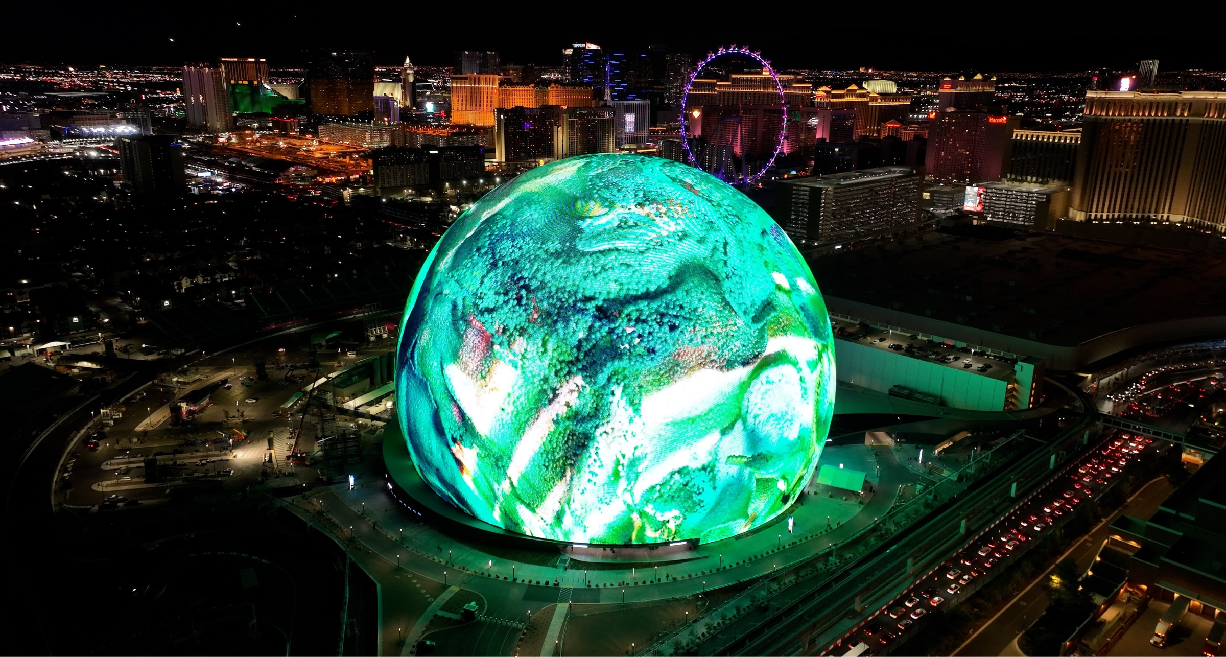 A stunning nighttime aerial view of the Las Vegas Sphere, illuminated with vibrant green and white digital visuals. The city skyline, including hotels, casinos, and the High Roller Ferris wheel, is visible in the background, showcasing the bright lights of the Las Vegas Strip. The futuristic display on the Sphere highlights cutting-edge technology and immersive digital experiences.