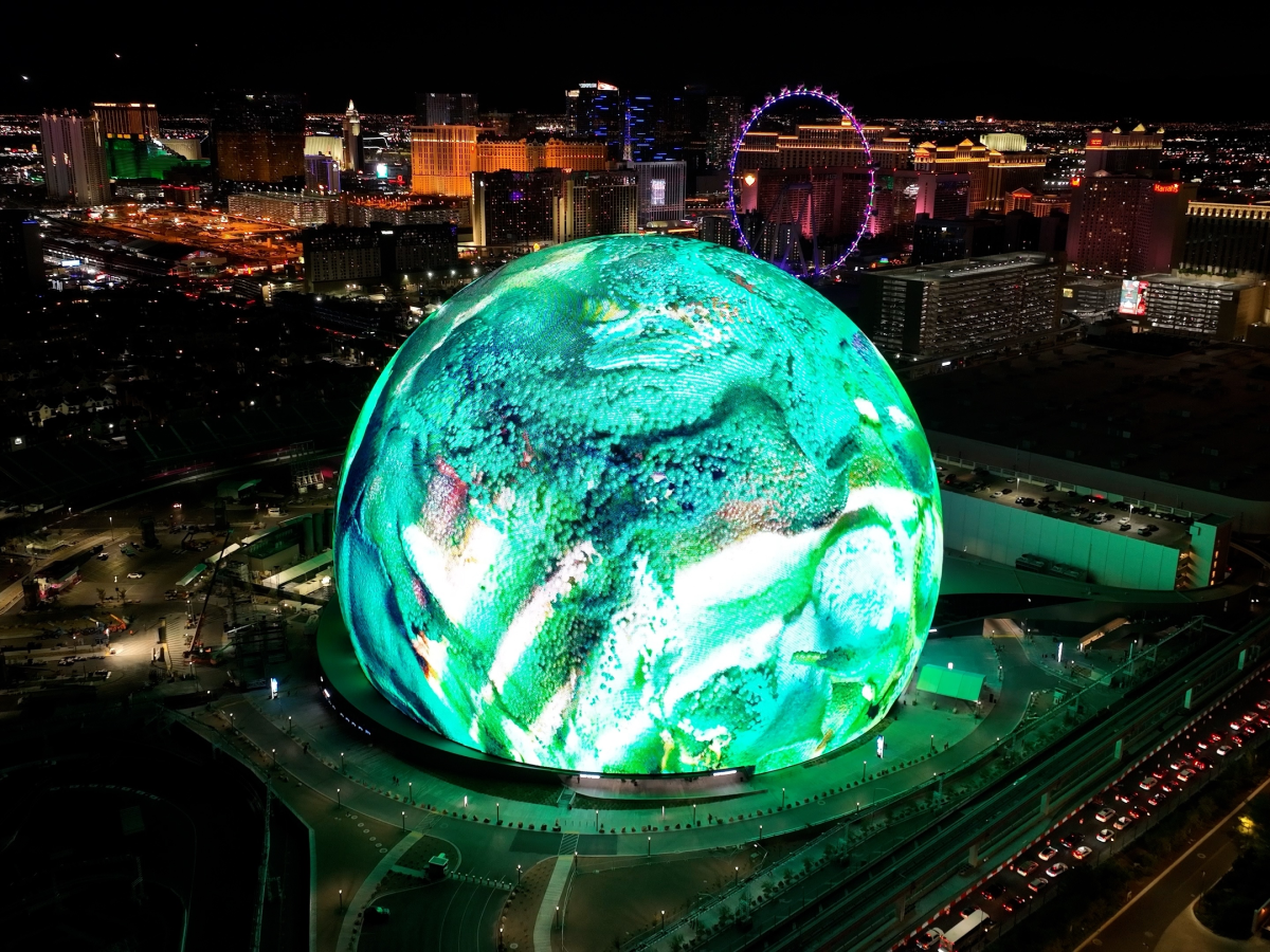 A stunning nighttime aerial view of the Las Vegas Sphere, illuminated with vibrant green and white digital visuals. The city skyline, including hotels, casinos, and the High Roller Ferris wheel, is visible in the background, showcasing the bright lights of the Las Vegas Strip. The futuristic display on the Sphere highlights cutting-edge technology and immersive digital experiences.
