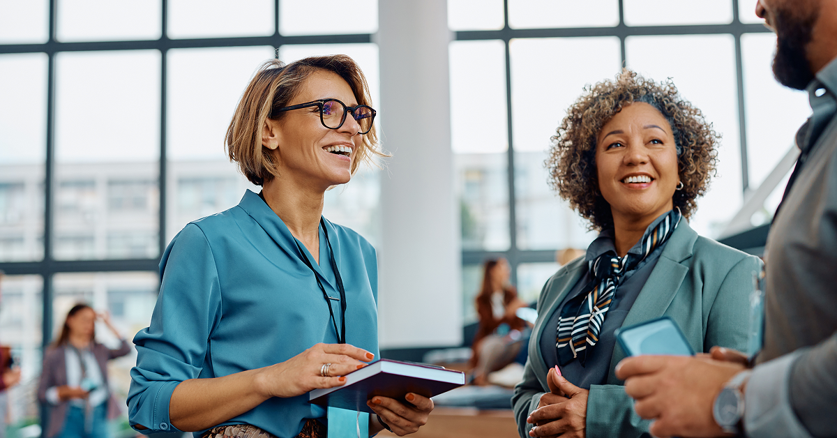 Three professionals engaging in a lively conversation at a networking event, with two women smiling and holding a notebook and phone, and a modern office background with large windows.