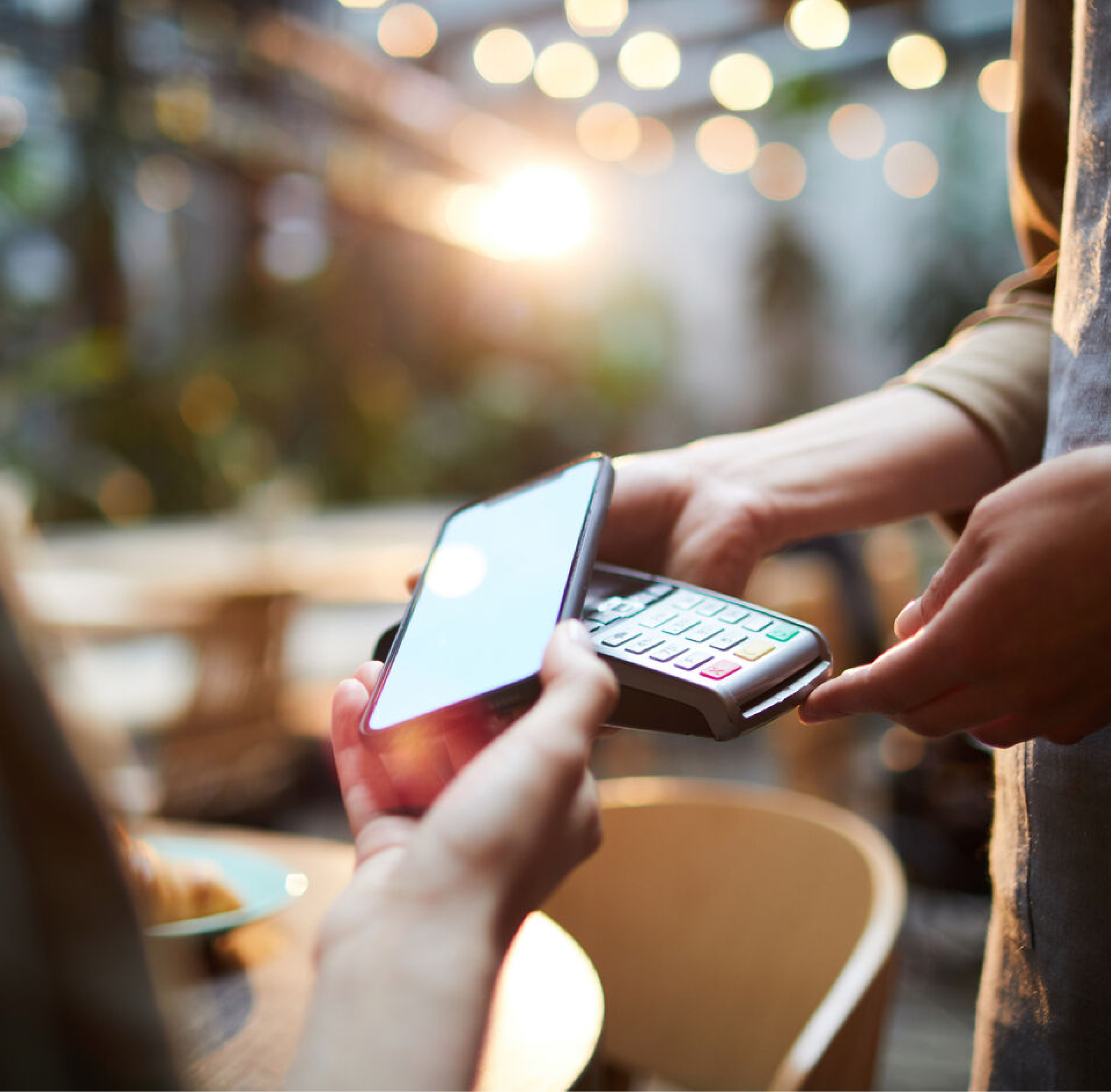 A close-up of a person making a contactless payment with a smartphone on a card reader, set against a blurred background with warm lighting and bokeh effects.