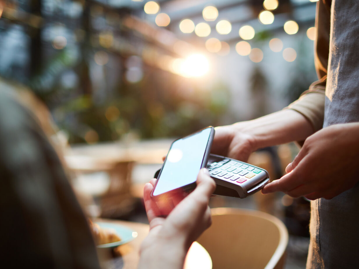 A close-up of a person making a contactless payment with a smartphone on a card reader, set against a blurred background with warm lighting and bokeh effects.