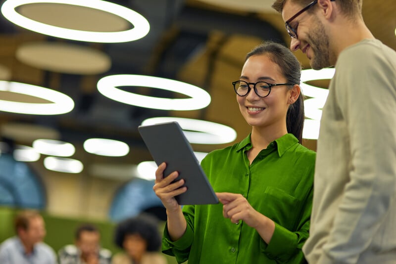 A smiling woman in a green blouse holds a tablet while standing next to a man in glasses and a light sweater, both looking at the screen together in a modern office space with circular overhead lights and a blurred group working in the background.